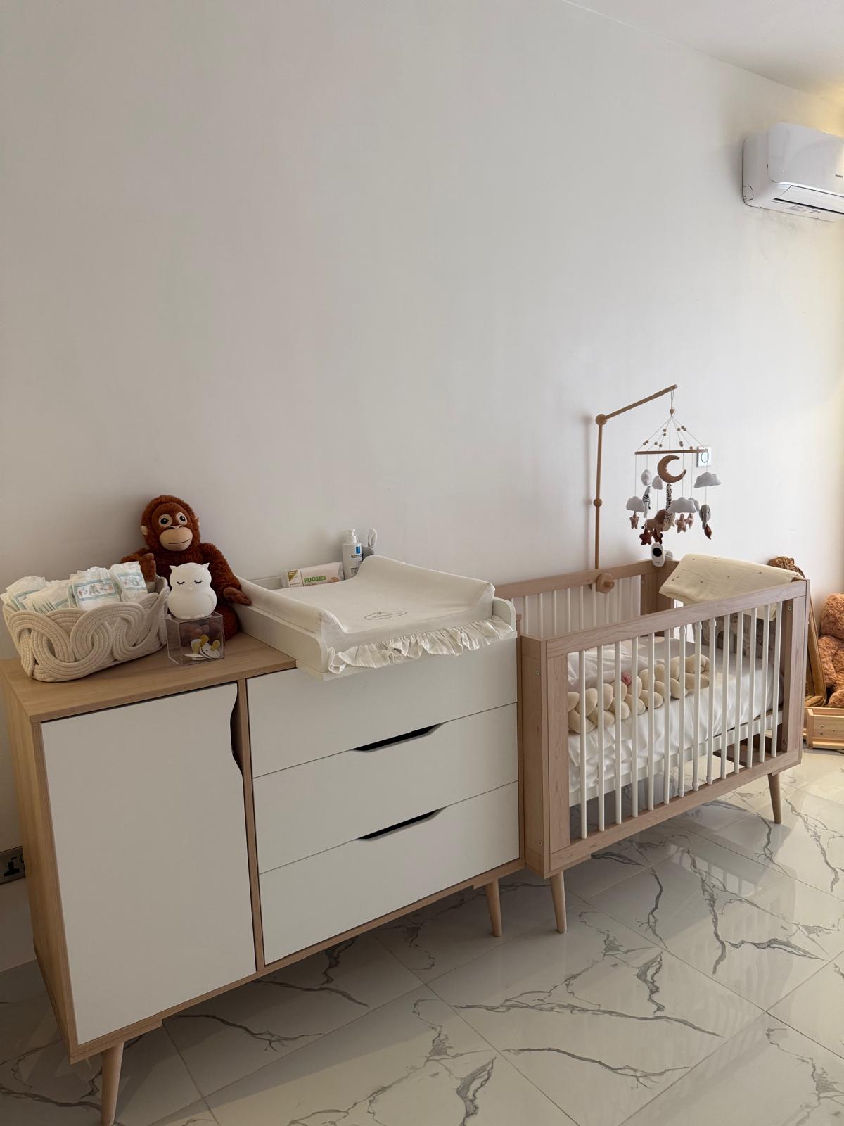 A mid-century modern baby Sofie furniture set featuring a changing table with a built-in changing pad and a light wood crib with a hanging mobile, set against a white wall on a marble-tiled floor in Lagos Nigeria