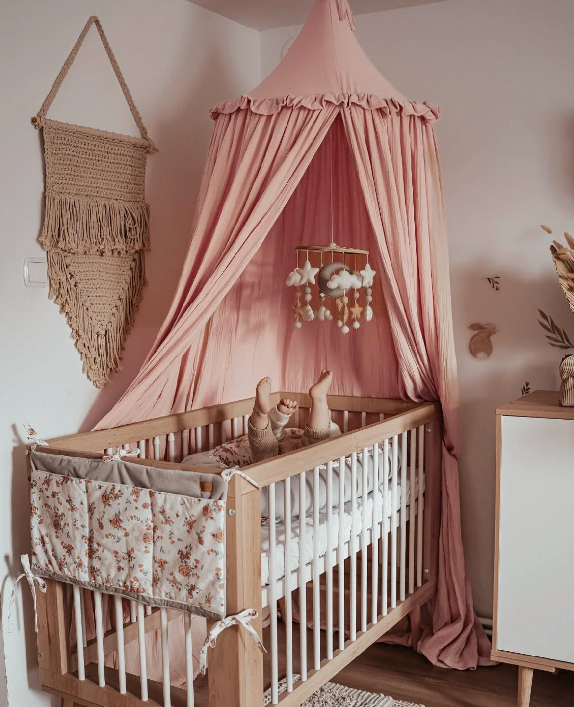 A pink canopy bed with Sofie wooden crib, floral bedding, and a hanging mobile in a softly decorated nursery in Lagos Nigeria 