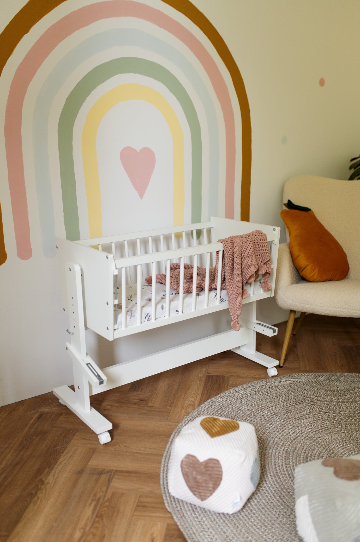 A white baby crib with a pink blanket draped over the side, set in a nursery with a pastel rainbow wall mural, wood floors, and a beige armchair.